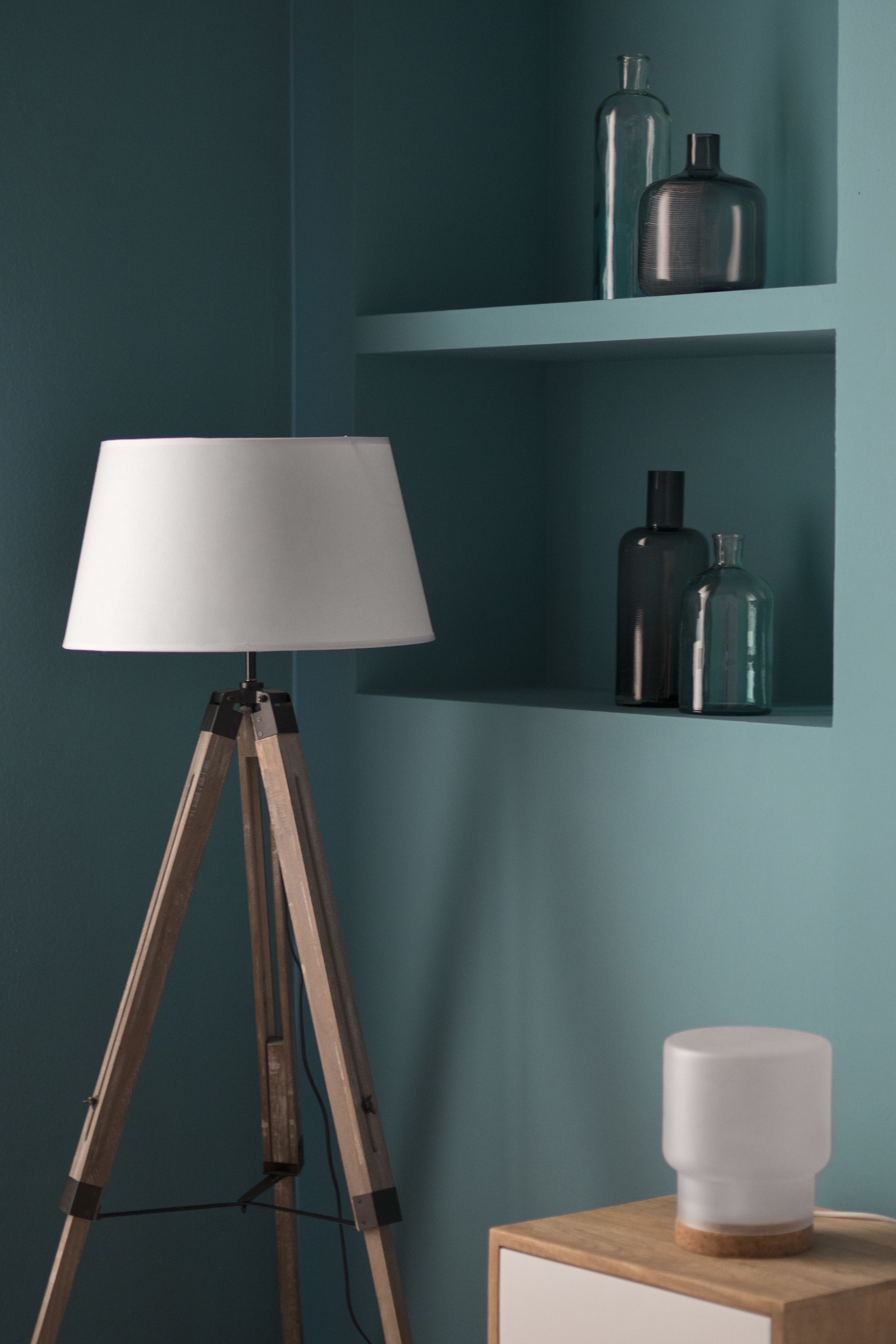 A tripod floor lamp with a white lampshade, a wooden nightstand with a white object, and teal-colored shelves with glass bottles.