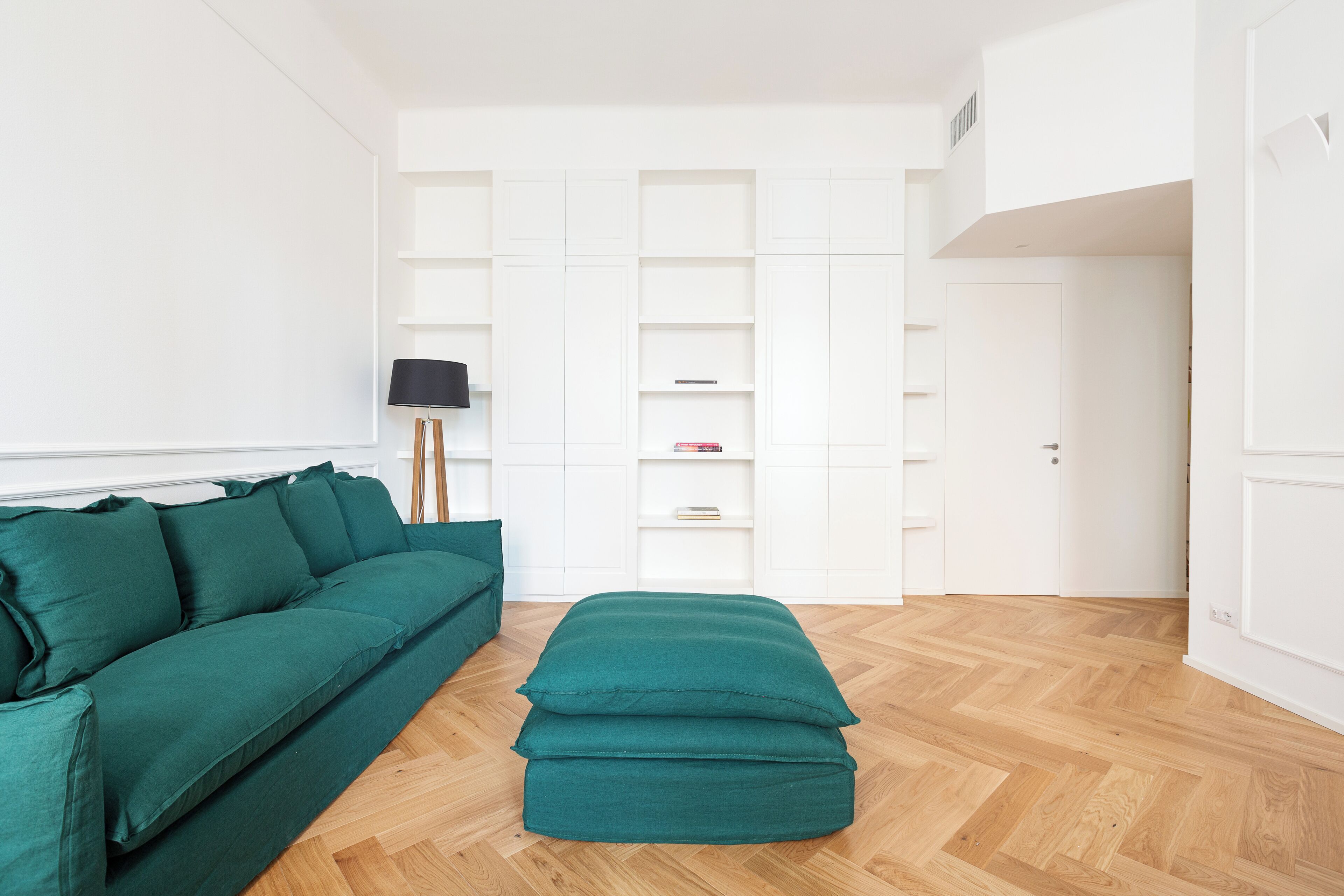 Ein modernes Wohnzimmer mit einem grünen Sofa, passendem Hocker und einer Stehlampe. An der Wand befindet sich ein weißes Regalsystem und rechts eine Tür.