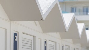 Een rij witte hotelkamers aan het strand met luiken, genummerd 3 en 4, en een bord met de tekst 'LUXOR BEACH' op de achtergrond.