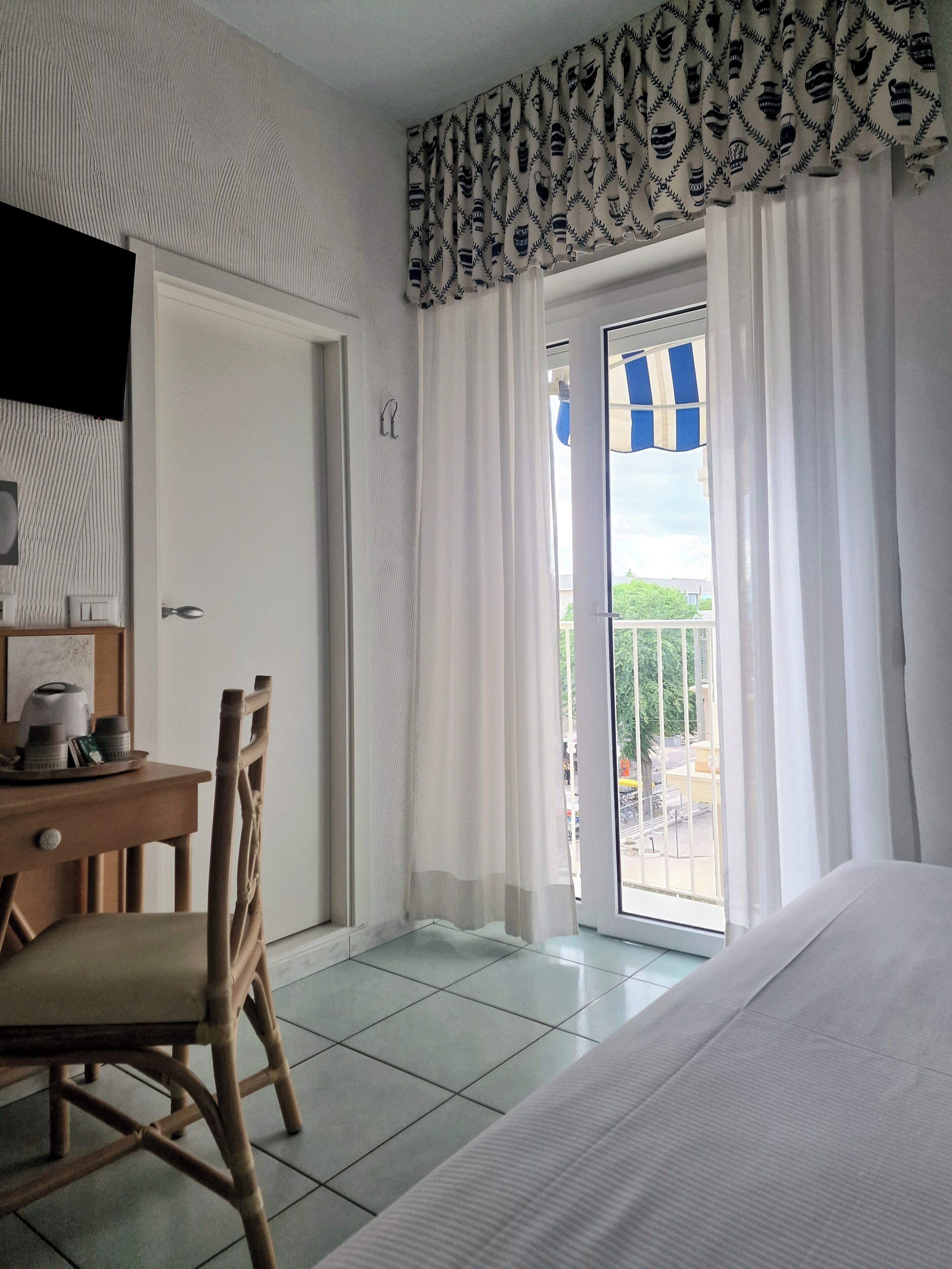 A hotel room with a bed, a wooden chair, a small table with a kettle and cups, and a view of the outside through a window with curtains.