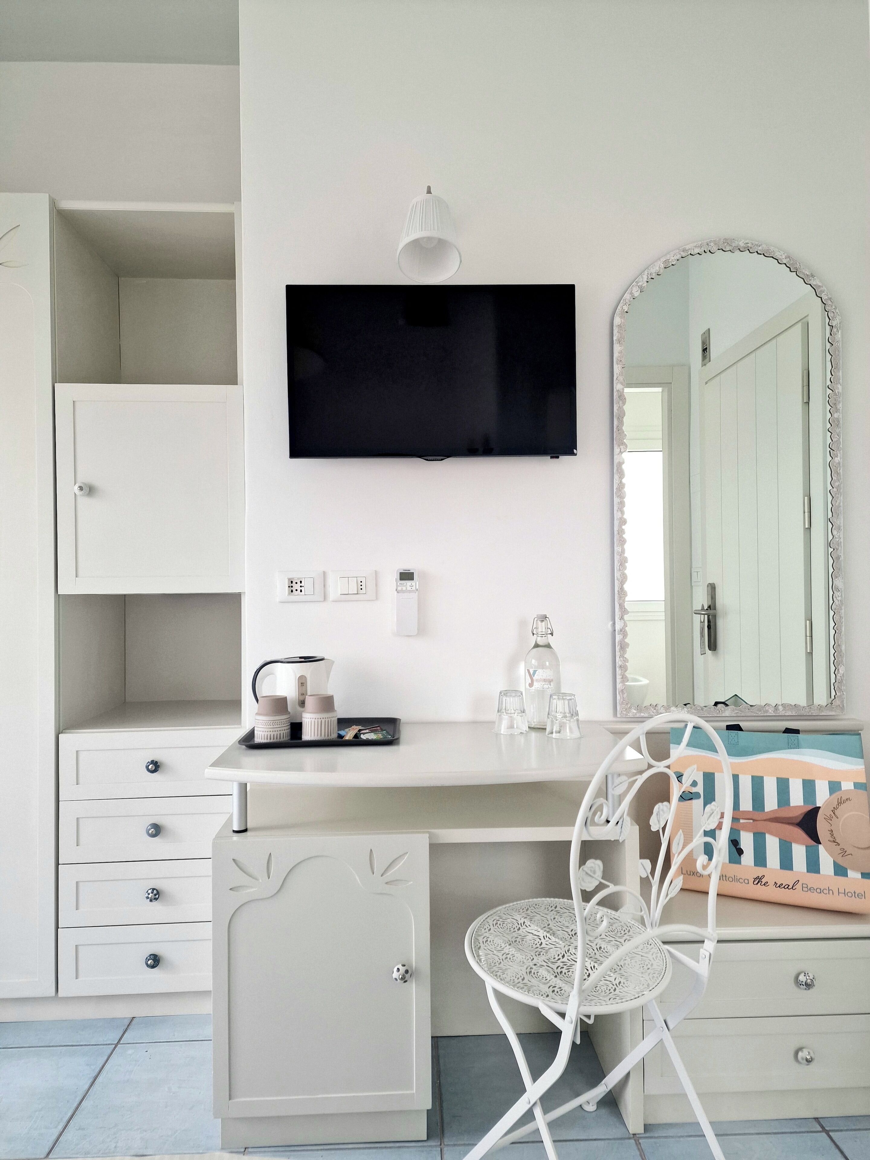 A white room with a TV mounted on the wall, a vanity with a mirror, and a chair with a decorative cushion.
