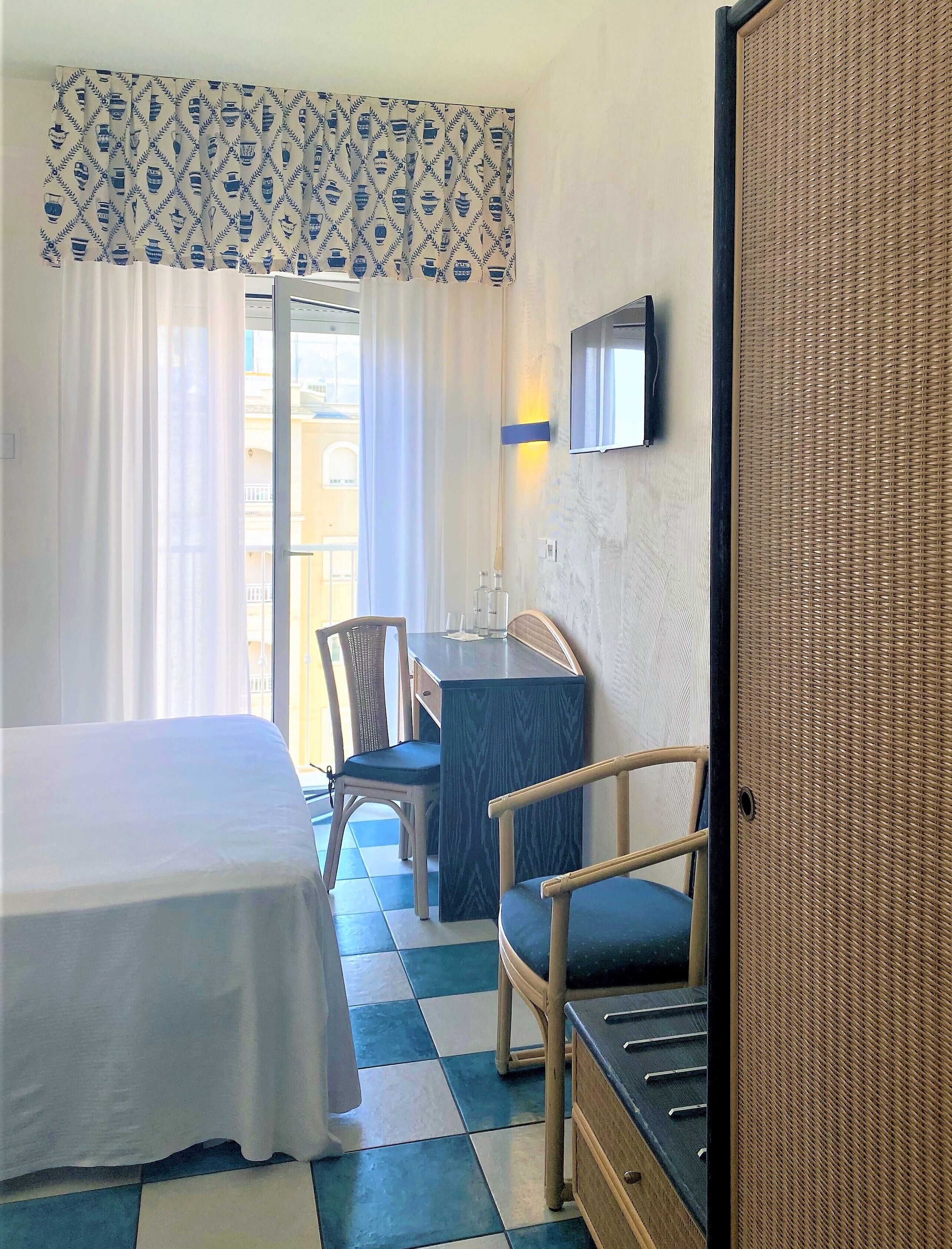 A hotel room with a bed, a desk with a chair, a TV mounted on the wall, and a window with curtains.