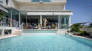 Een zwembad met een glazen balustrade, een overdekt terras en een modern gebouw met grote ramen.