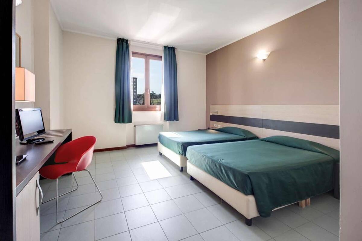 Hotellihuone, jossa on kaksi sänkyä, työpöytä tietokoneella, punainen tuoli ja ikkuna, jossa on verhot.