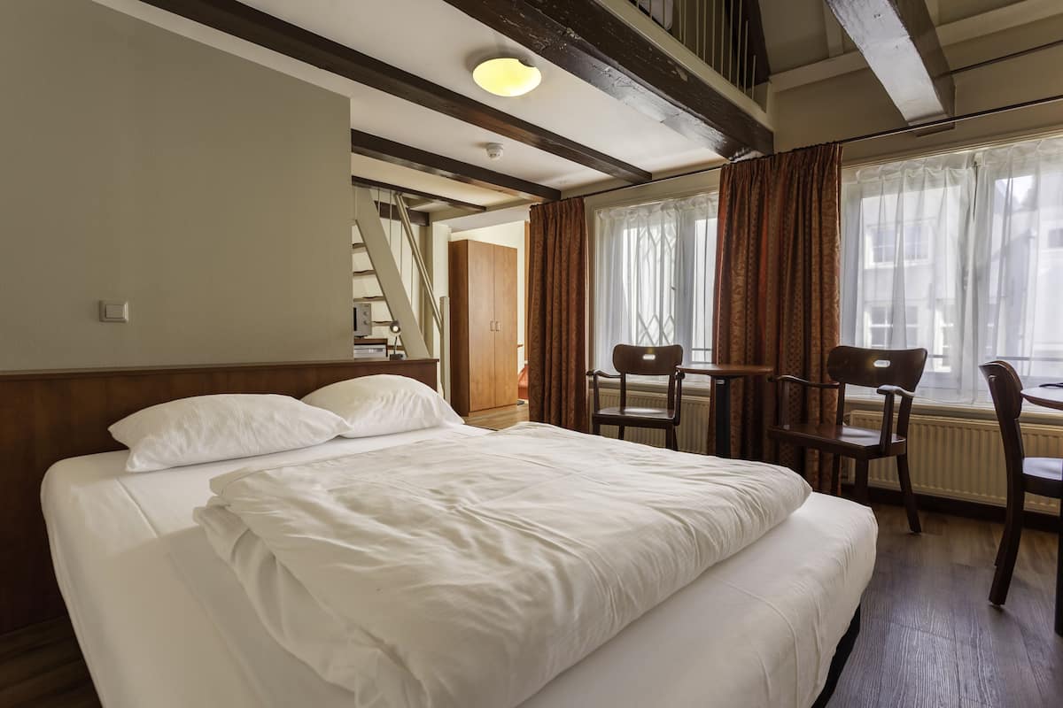 Quarto de hotel com uma cama grande, cabeceira de madeira, uma escrivaninha com duas cadeiras e uma janela com cortinas.