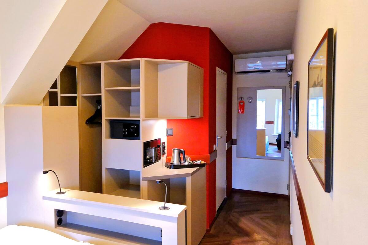 Quarto de hotel com uma secretária branca, uma parede em destaque vermelha, uma estante embutida, uma máquina de café e um espelho.