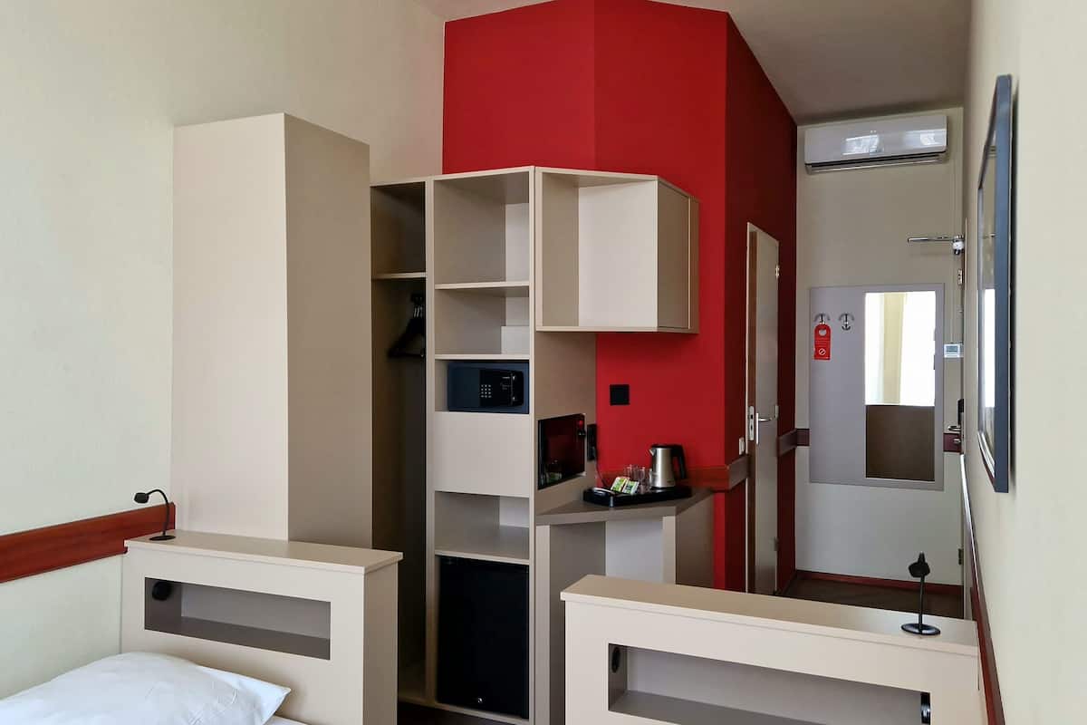 Quarto de hotel com cama, guarda-roupa, secretária com espelho, parede de destaque vermelha e cozinha compacta com cafeteira.