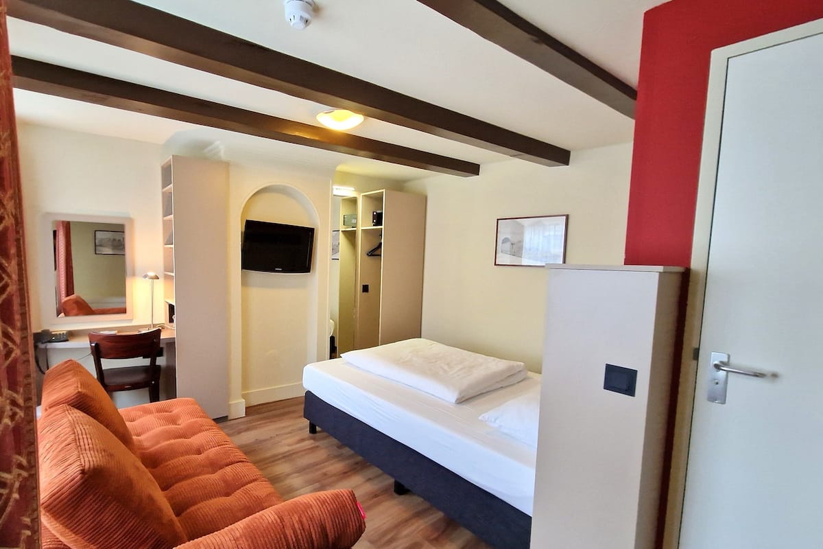 Quarto de hotel com cama, sofá, escrivaninha com cadeira, televisão e guarda-roupa.