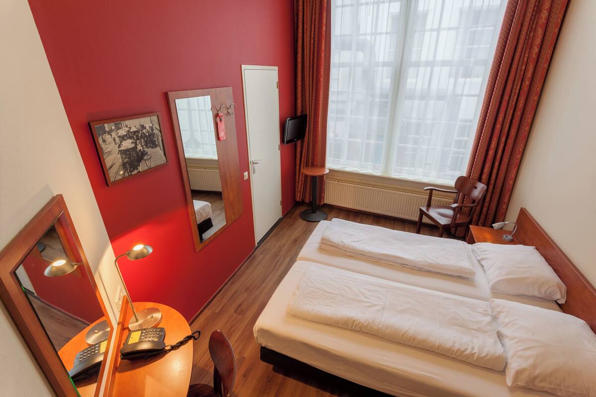 Quarto de hotel com duas camas, uma escrivaninha com telefone, uma cadeira, um espelho e uma janela com cortinas.