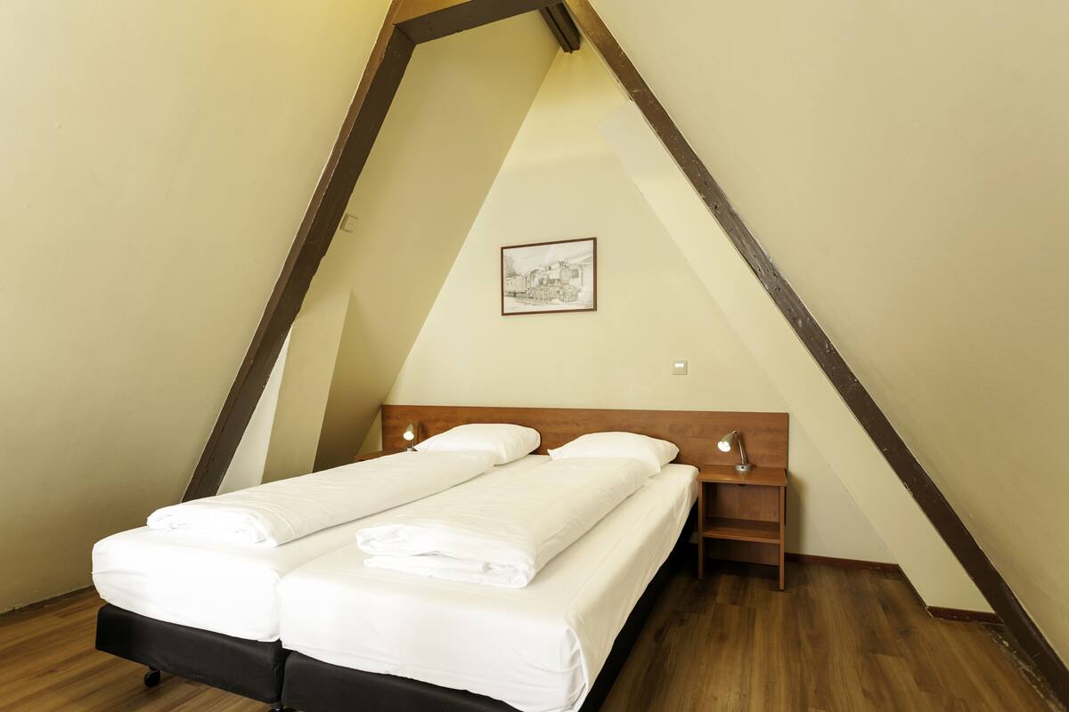 Quarto com teto inclinado, uma cama com roupa de cama branca, mesa de cabeceira com abajur e um quadro na parede.
