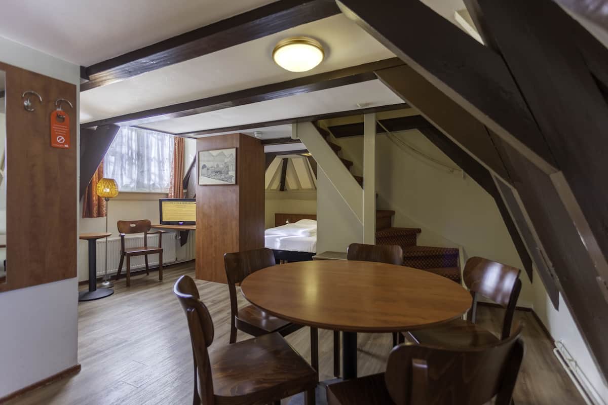 Uma área de refeições com uma mesa redonda de madeira e cadeiras, um pequeno espaço de cozinha com um frigorífico e uma pia, e uma escada que leva a um andar superior.