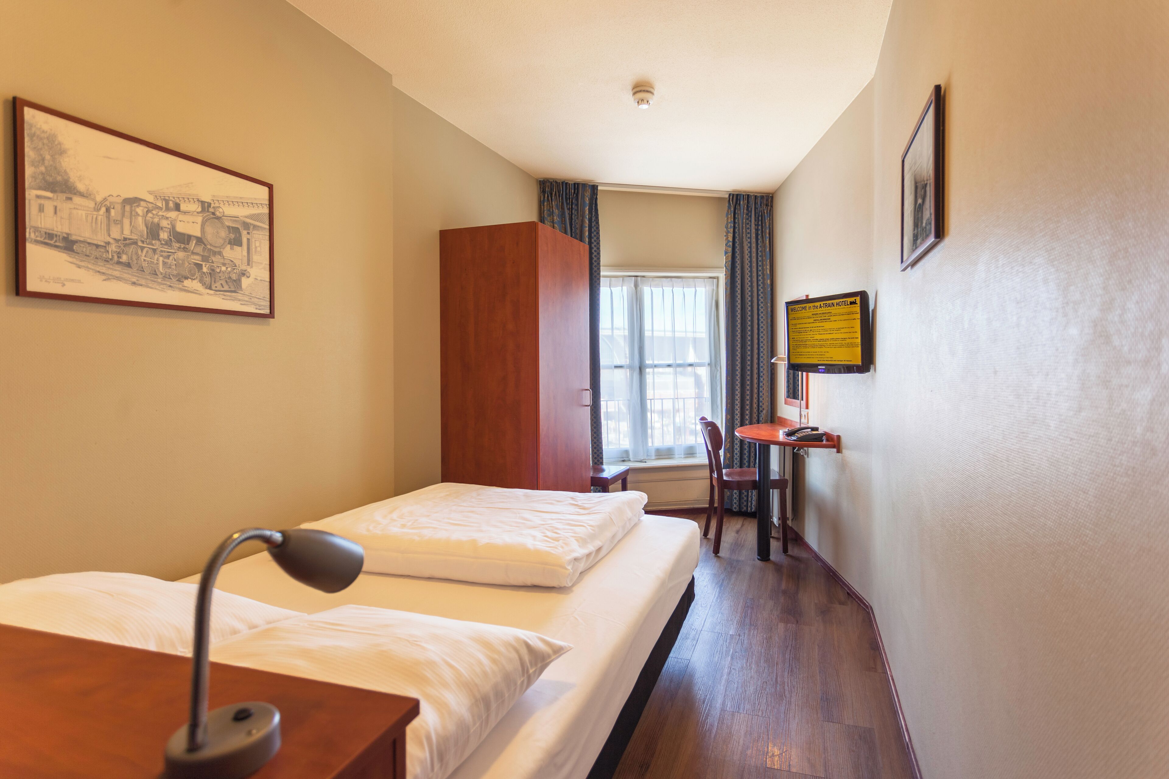 A hotel room with a bed, a desk, a chair, a lamp, a window, curtains, and a framed picture on the wall.