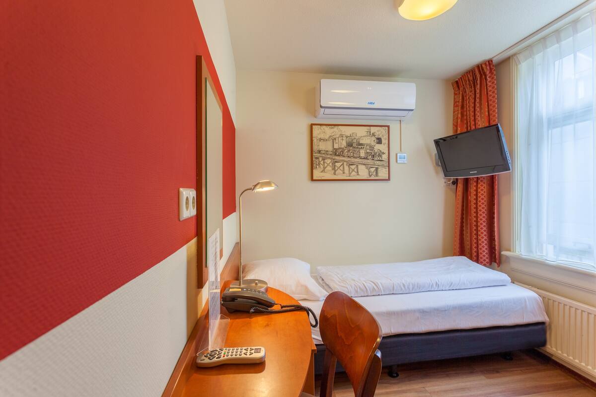 Quarto de hotel com cama, escrivaninha com telefone e comando remoto, ar-condicionado na parede, televisão e janela com cortinas.