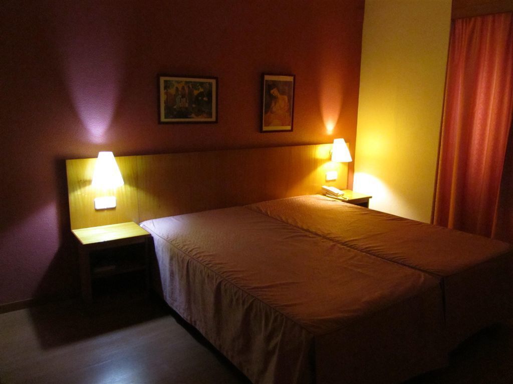Quarto de hotel com cama, duas luminárias de cabeceira e duas imagens emolduradas na parede.