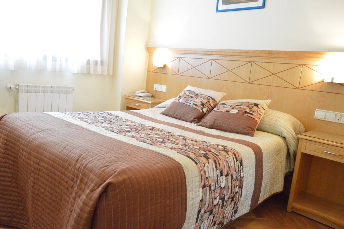 A neatly made bed with a patterned bedspread, two bedside tables with lamps, a radiator, and a framed picture on the wall.