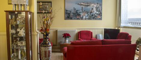 Una sala de estar con un sofá rojo, un aparador de vidrio con adornos, un florero con flores y una pintura de un paisaje urbano en la pared.
