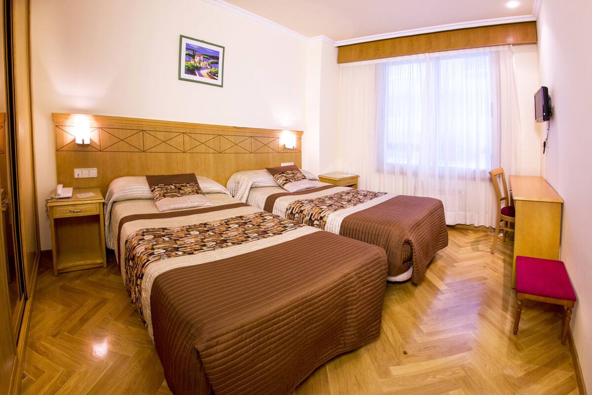 A hotel room with two beds, wooden headboard, bedside tables with lamps, a desk with a chair, and a window with curtains.