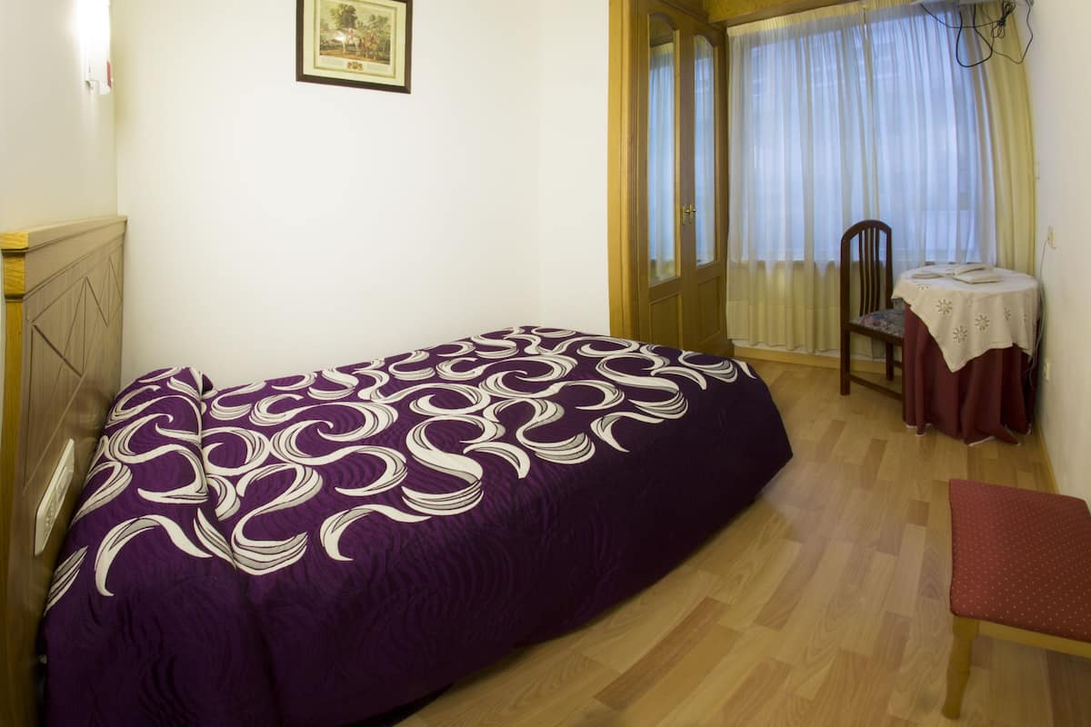 A room with a bed, a chair, a table with a tablecloth, a picture on the wall, and a window with curtains.