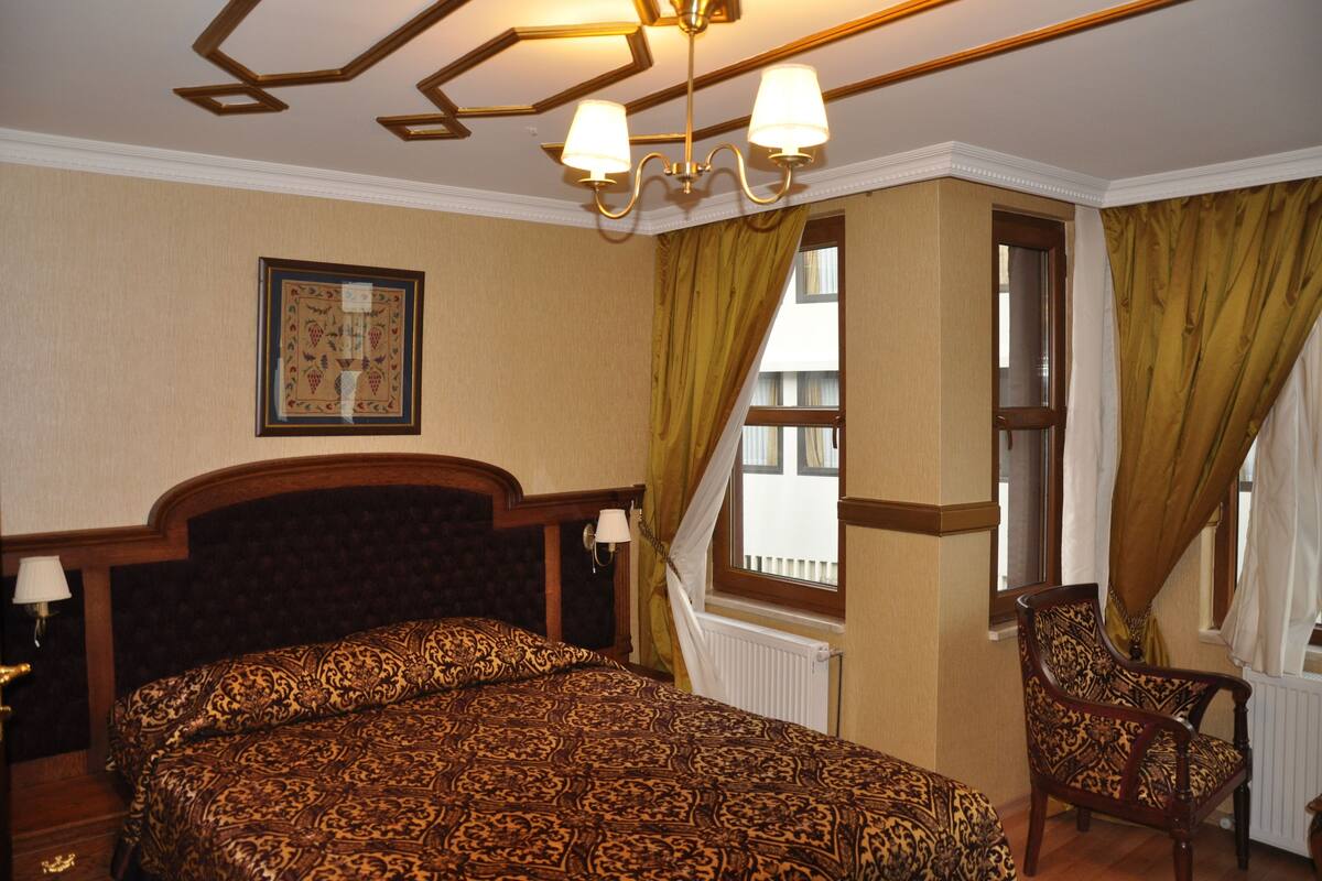 Une chambre avec un grand lit, une chaise, une fenêtre avec des rideaux, un lustre et un tableau encadré au mur.