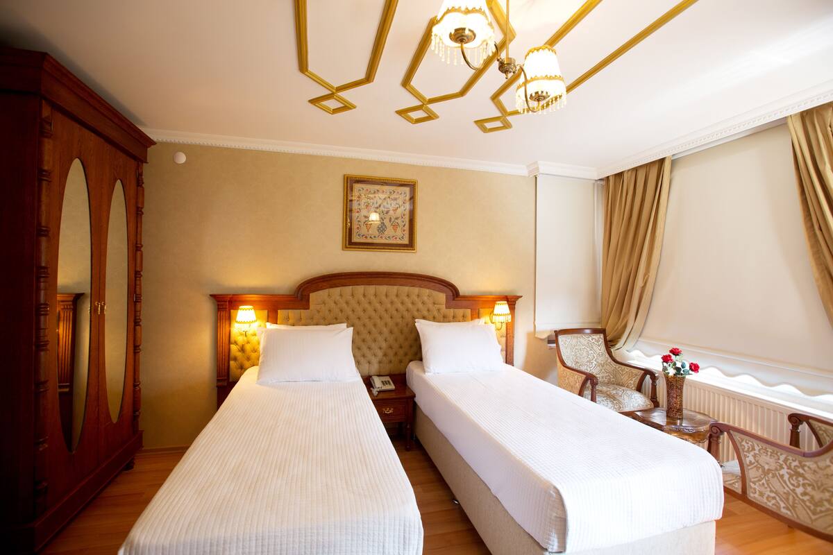 Une chambre d’hôtel avec deux lits simples, une armoire en bois, un lustre, un tableau encadré au mur et une fenêtre avec des rideaux.