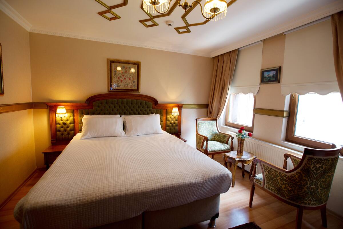 Une chambre d’hôtel avec un grand lit, une tête de lit verte, deux lampes de chevet, une petite table avec un vase de fleurs, un fauteuil vert et une fenêtre avec des rideaux.