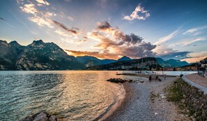 A lakeside view with mountains in the background, a pebble beach, and a marina with boats. - Hotel Residence Torbole (Nago-Torbole)