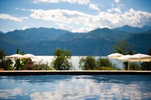 Piscina con vistas a montañas y un lago. Hay sombrillas y una persona de pie junto a la piscina.