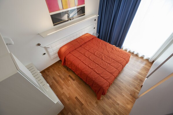 Een slaapkamer met een houten vloer, een bed met een oranje sprei, een wit hoofdeinde, een klein raam met blauwe gordijnen, en een wandplank boven het bed.