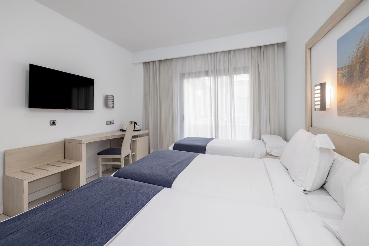 A hotel room with two beds, a desk, a chair, a TV, and a window with curtains.