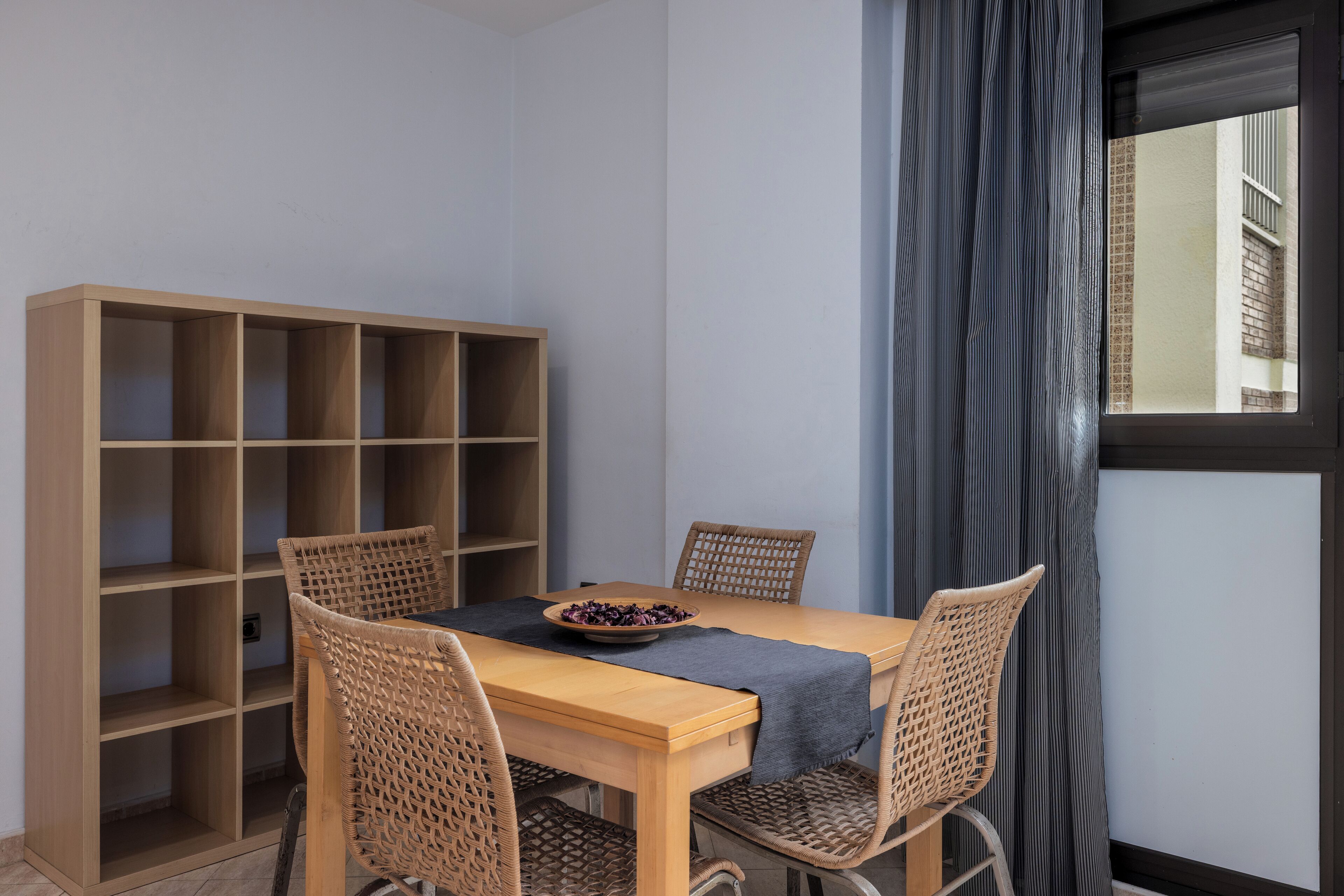 A dining area with a wooden table, four wicker chairs, a dark blue table runner, and a bookshelf with multiple shelves.