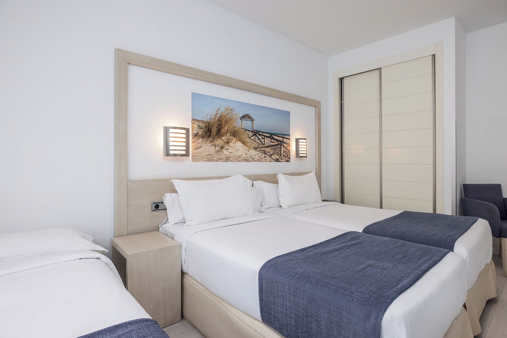Een hotelkamer met twee bedden, een groot gepresenteerd schilderij van een strand, en een schuifdeurkast.