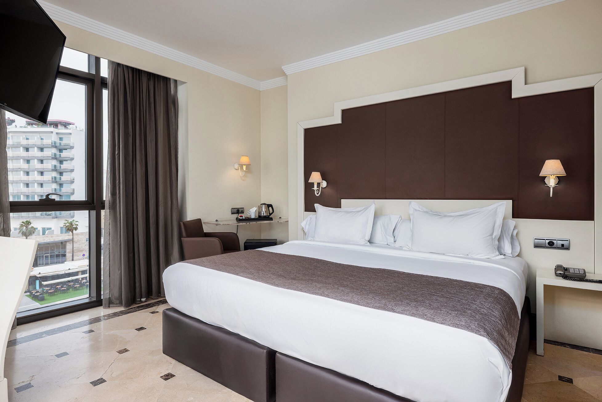 A hotel room with a large bed, a desk, two bedside lamps, and a view of the city through large windows.