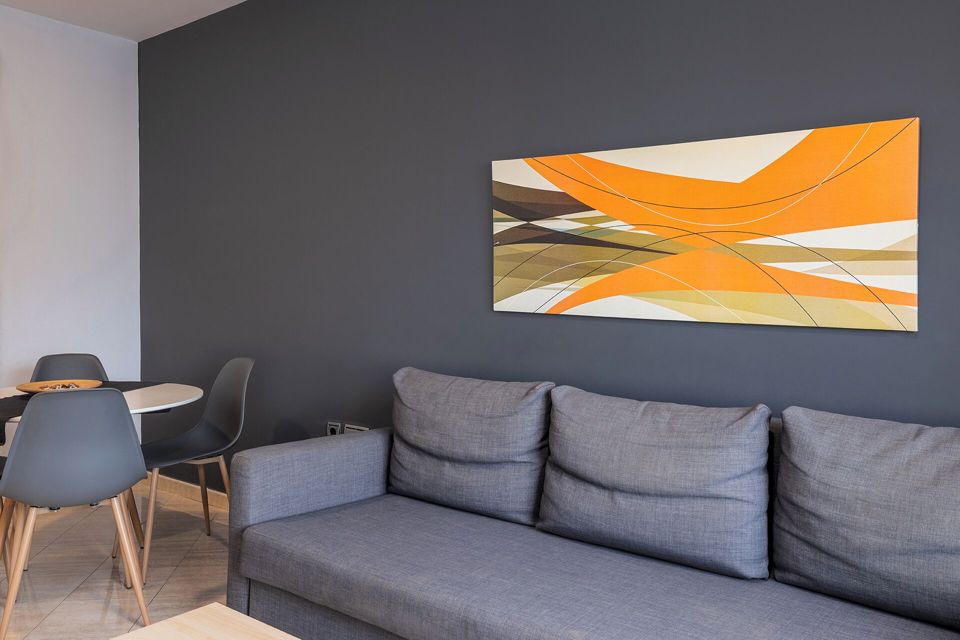 A modern living room with a grey sofa, a small round table, and two chairs. A colorful abstract painting is on the wall.