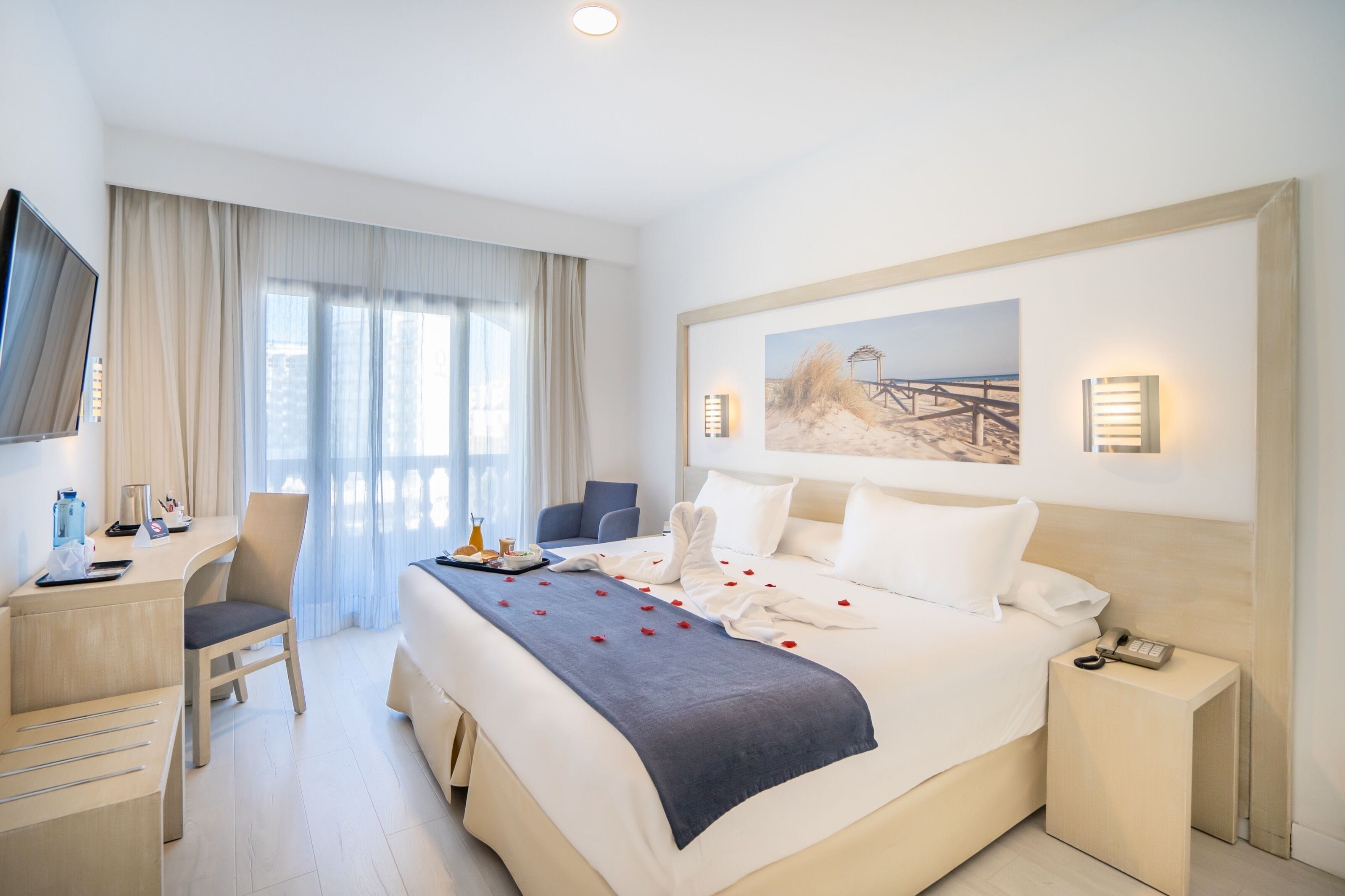 A hotel room with a bed, a desk, a chair, a TV, and a painting on the wall.