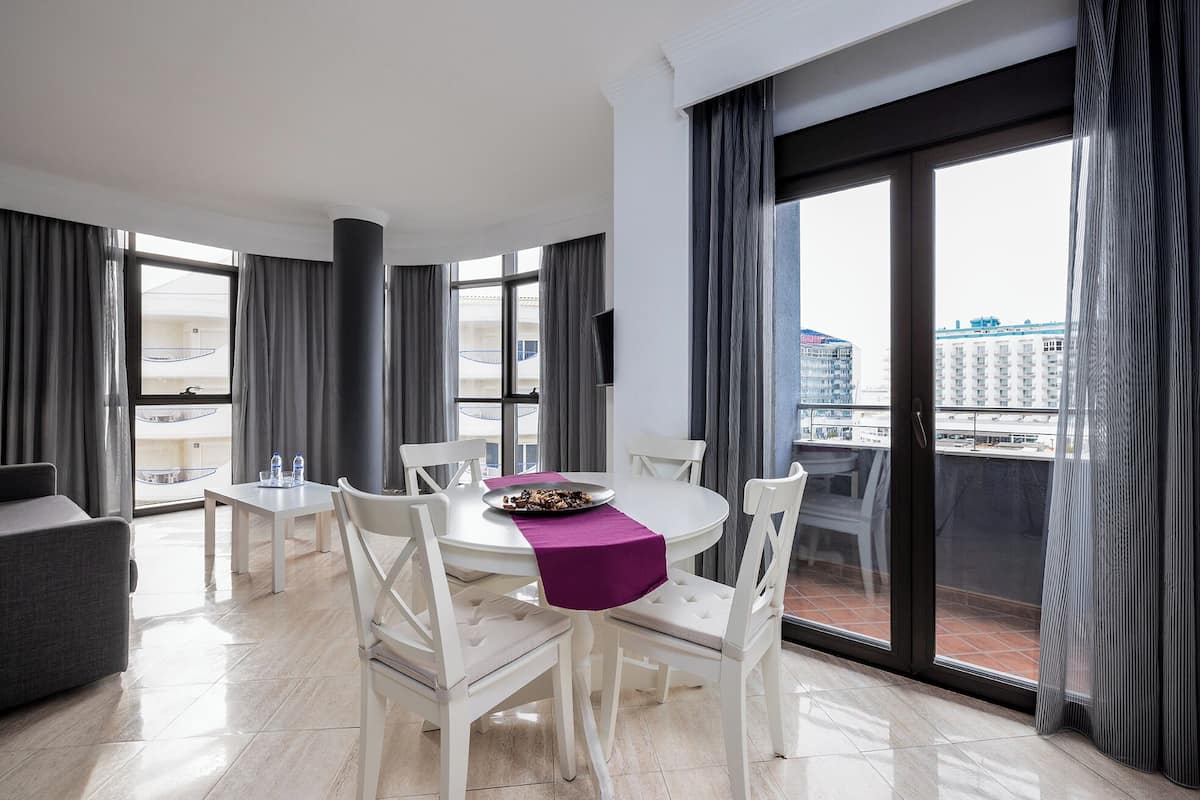 A modern hotel room with a dining area, large windows, and a view of the cityscape.