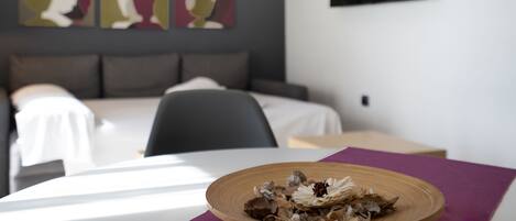 Hotellihuone, jossa on sänky, yöpöytä ja koristeellinen lautanen violetilla pöytäliinalla katetulla pöydällä.