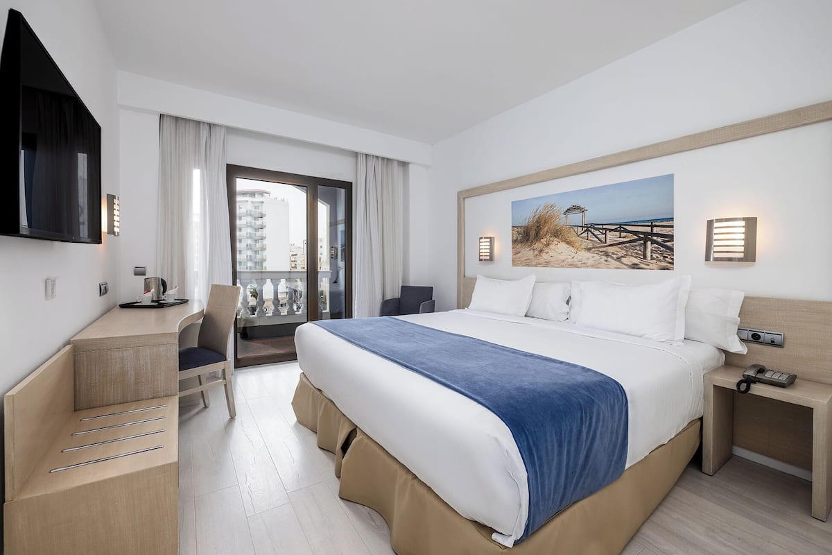 A hotel room with a bed, a desk, a chair, a TV, and a balcony with curtains.