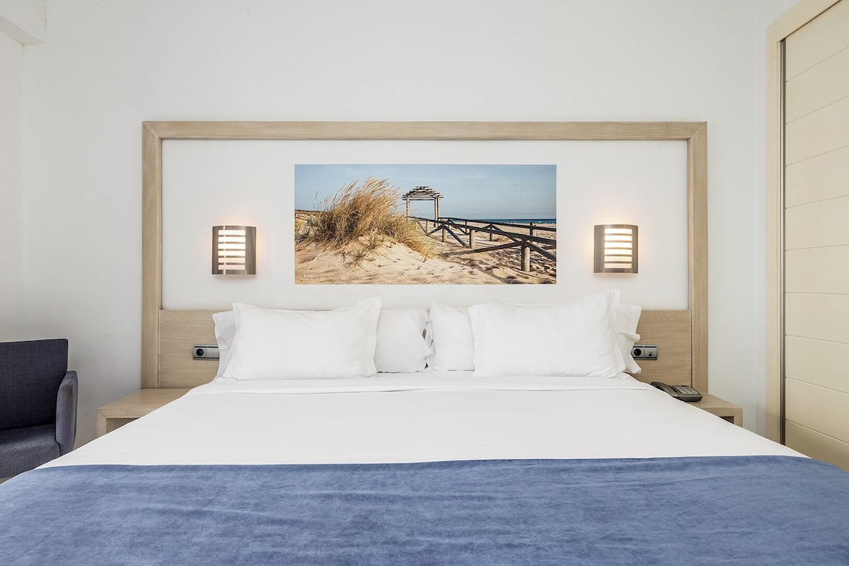 A hotel room with a bed, a headboard with a beach scene, two wall-mounted lights, and a nightstand with a phone.