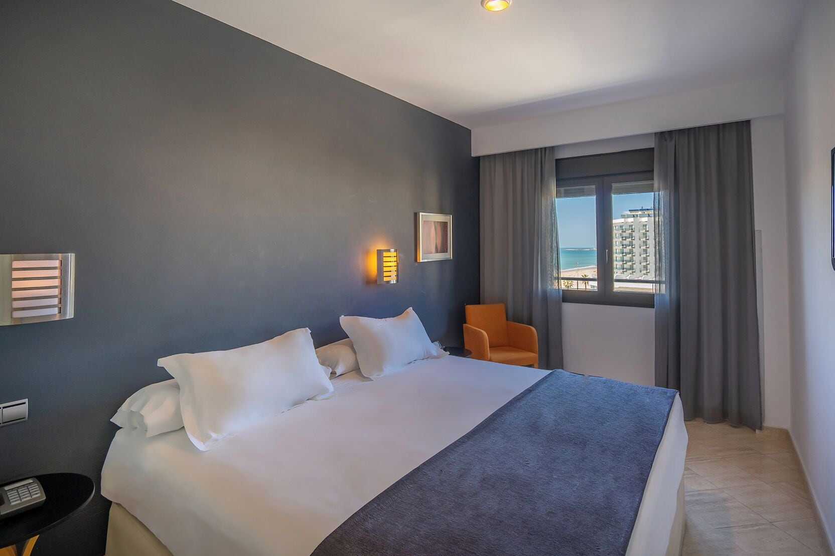 A hotel room with a large bed, a chair, a window with a view of the beach, and a wall-mounted light fixture.