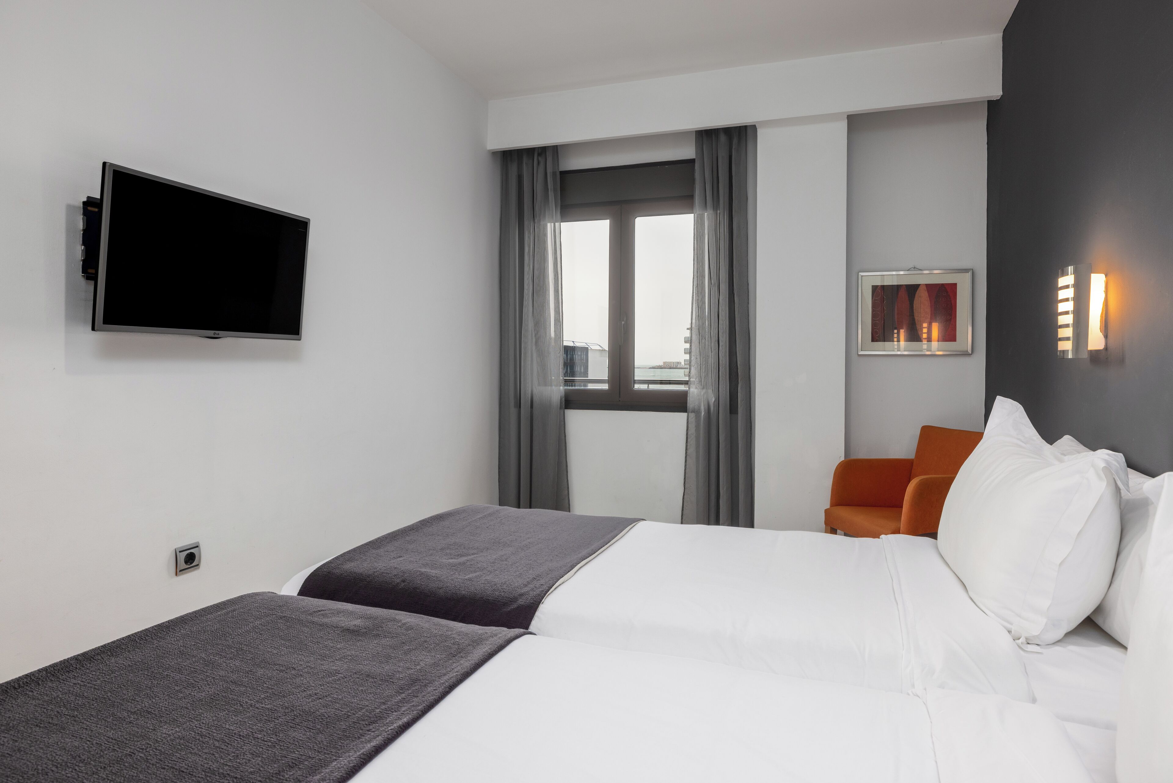 A hotel room with a bed, a TV mounted on the wall, a window with curtains, and a chair.
