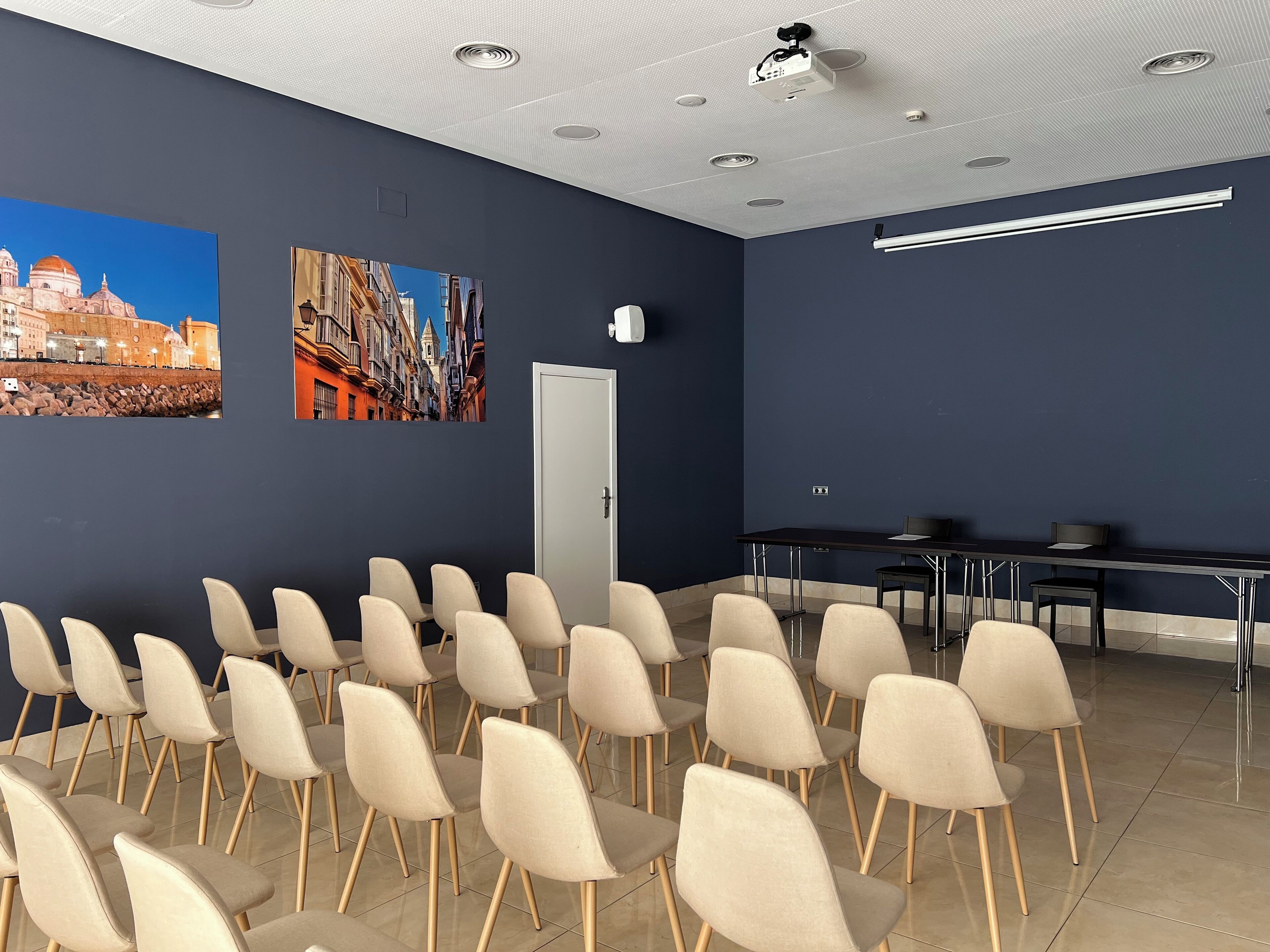 A conference room with rows of beige chairs, a projector, and two large wall-mounted pictures depicting cityscapes.
