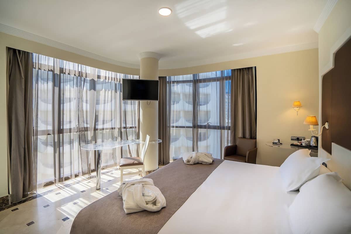 A hotel room with a large bed, a desk, a chair, a TV, and a view through the windows.