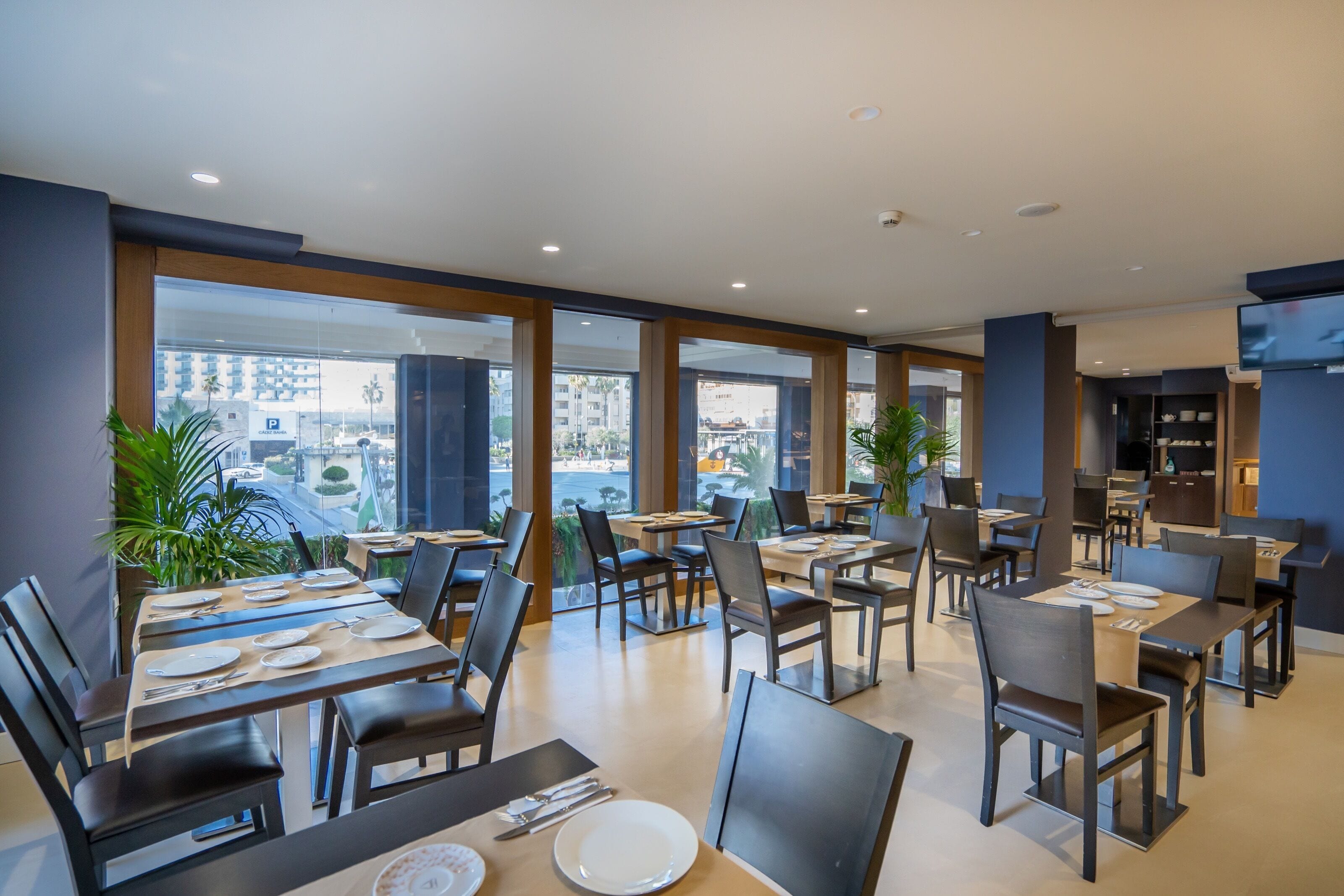 A modern restaurant with tables set for dining, large windows offering city views, and a flat-screen TV mounted on the wall.