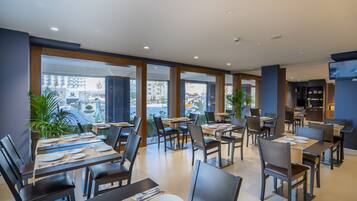 Un restaurante moderno con mesas dispuestas para comer, amplios ventanales con vistas a la ciudad y un televisor de pantalla plana en la pared.