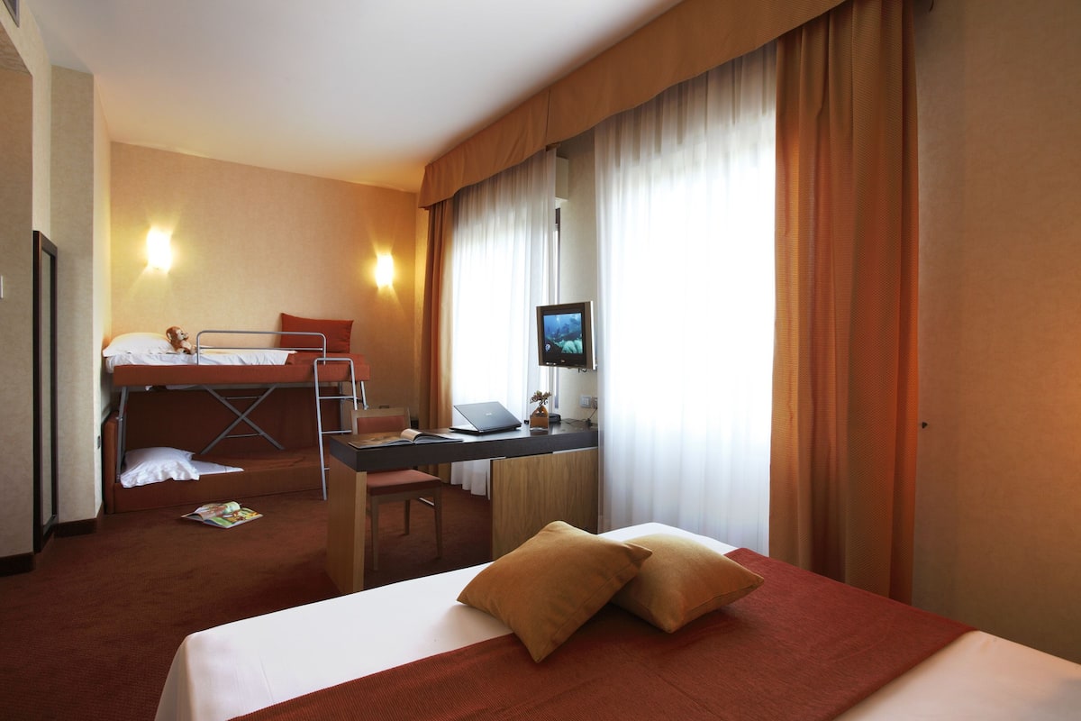 A hotel room with a bunk bed, a desk with a laptop, a TV, and a bed with two pillows.