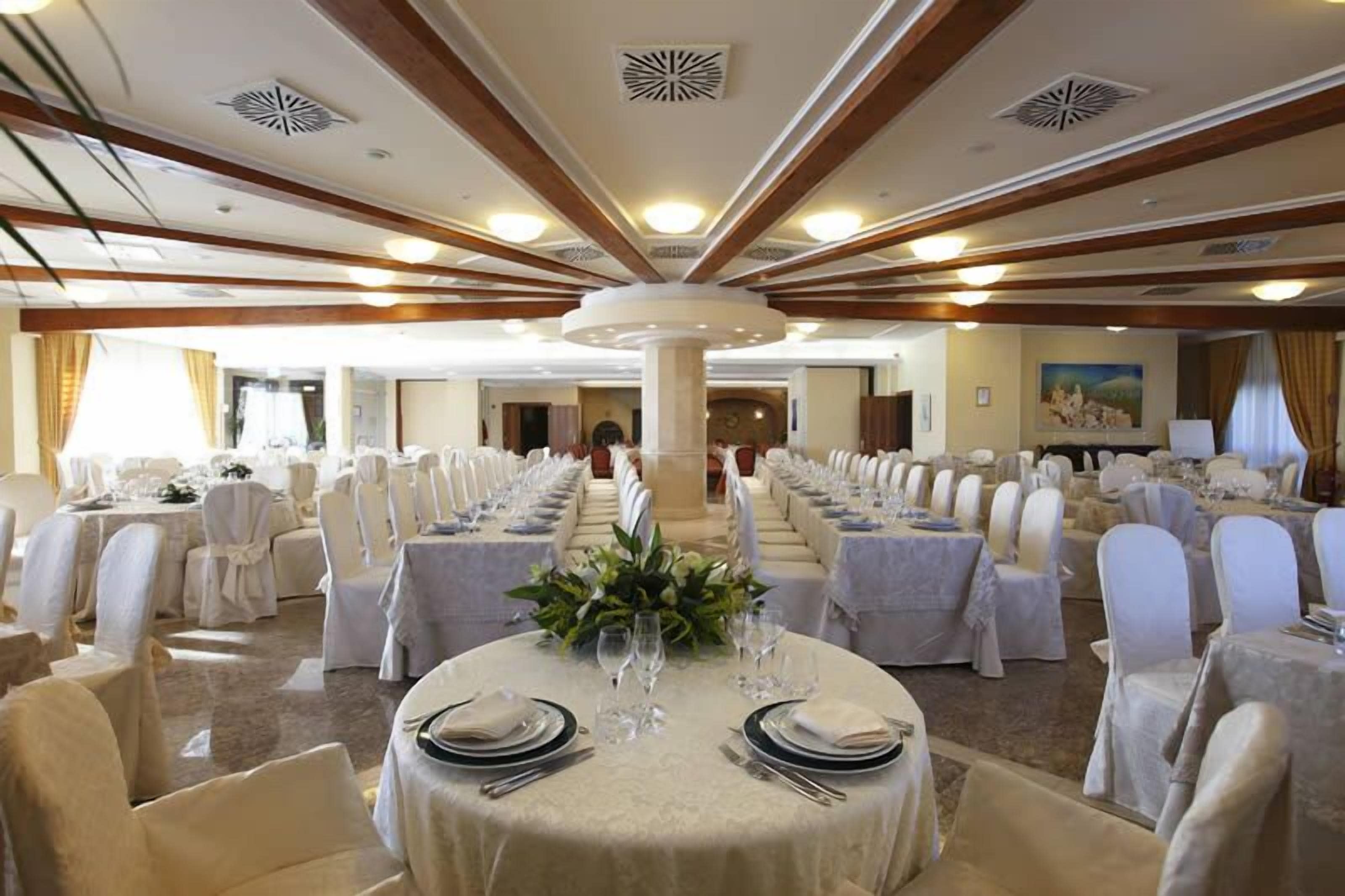 Una sala banchetti con tavoli rotondi apparecchiati per la cena, sedie bianche e una colonna centrale.