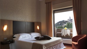 A hotel room with a bed, bedside tables, a chair, a balcony with a table and chairs, and a view of a hill with a building. - Villa Luisa (Todi)
