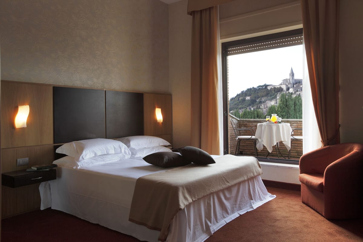 A hotel room with a bed, bedside tables, a chair, a balcony with a table and chairs, and a view of a hill with a building.