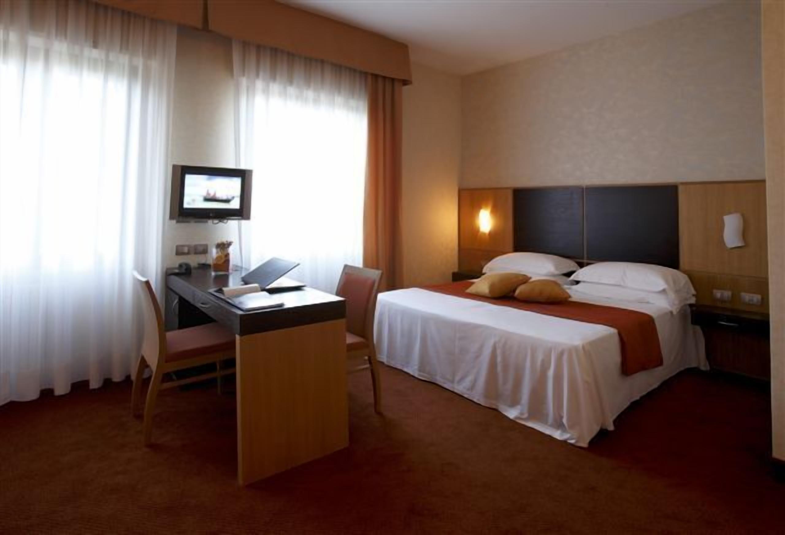 Una habitación de hotel con cama, escritorio, silla, televisor y ventana con cortinas.