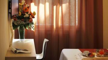 A hotel room with a dining table set for breakfast, a vase of flowers, and a TV mounted on the wall.